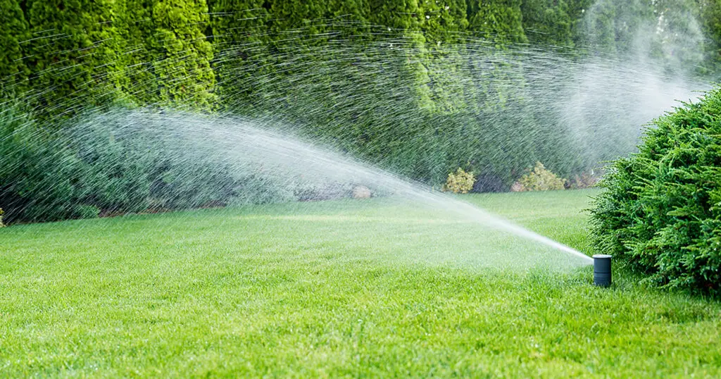 Smart Irrigation Systems - Image of new technology lawn irrigation systems with sprinklers spraying water on a beautiful landscaped lawn, with shrubs and a row of privacy trees.